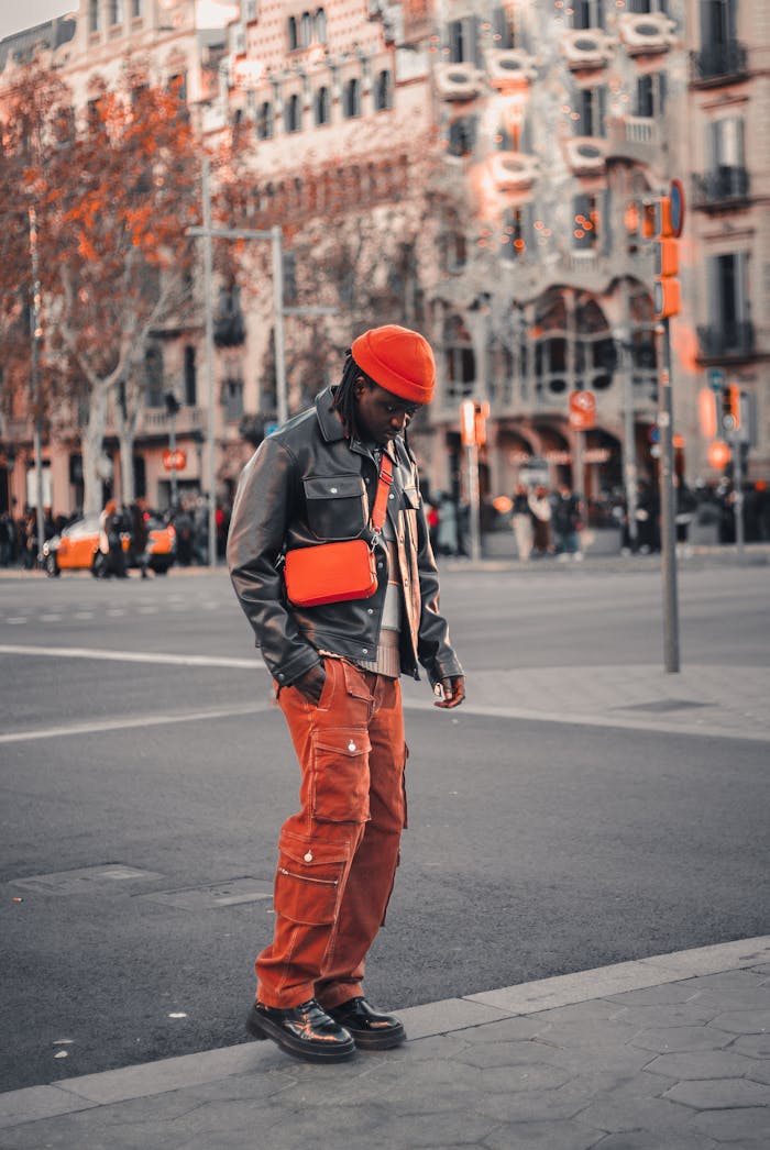 Stylish man in urban fashion, standing on city street, looking down with modern architecture backdrop.